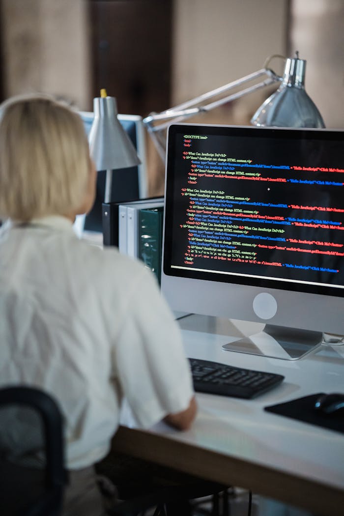 about-us Professional woman coding at office desk with two monitors and desk lamps.